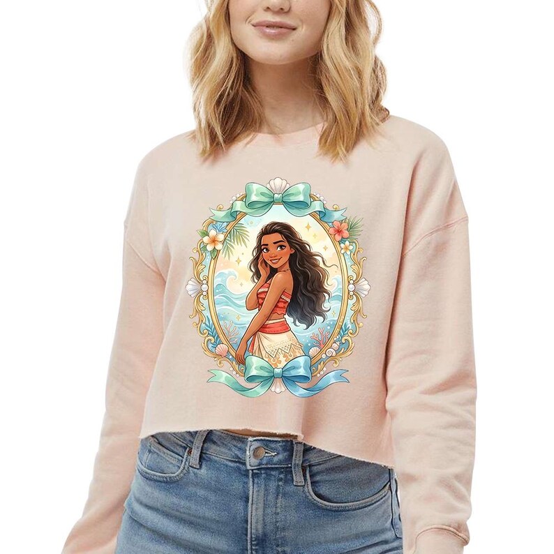 Peut inclure: Un sweat-shirt court rose clair avec une illustration de dessin anim&eacute; encadr&eacute;e d'une femme aux longs cheveux noirs, v&ecirc;tue d'une tenue rouge et blanche. Le cadre comprend des n&oelig;uds bleus, des fleurs et une sc&egrave;ne oc&eacute;anique tropicale. Le sweat-shirt est associ&eacute; &agrave; un jean bleu.