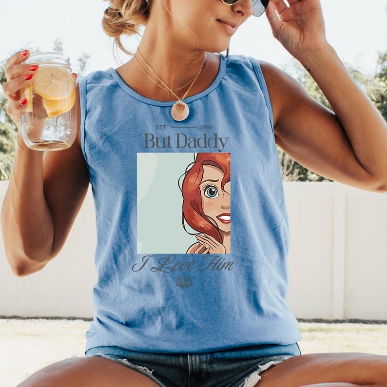 May include: Light blue tank top with the text "But Daddy" and "I Love Him". The graphic features a cartoon image of a character with red hair and blue eyes. The tank top is made of a soft material.