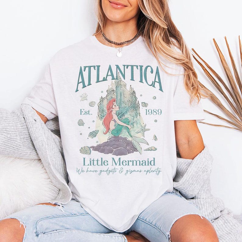 May include: White t-shirt featuring a graphic of the Little Mermaid. The design includes the text "Atlantica" above a castle illustration, with "Est. 1989" and "Little Mermaid" below. The shirt also has the phrase "We have gadgets & gizmos aplenty."