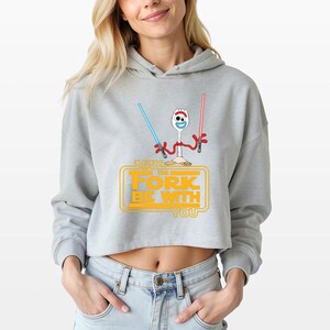 May include: A light gray cropped hoodie featuring a graphic of a white plastic spork character with red pipe cleaner arms holding a blue and a red lightsaber. Below the character, yellow text reads MAY THE FORK BE WITH YOU in a stylized font.