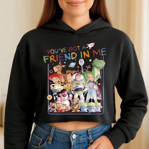 Może przedstawiać: Czarna kr&oacute;tka bluza z kapturem z kolorową grafiką postaci z filmu "Toy Story". Grafika zawiera tekst "YOU'VE GOT A FRIEND IN ME" w tęczowej czcionce, a także wizerunki Woody'ego, Buzza Astrala i innych postaci.