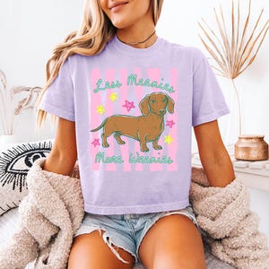 May include: A light purple short-sleeve t-shirt featuring a brown dachshund illustration in the centre. The graphic includes pink vertical stripes, colourful star accents, and the green text "Less Meanies More Weenies" printed above and below.