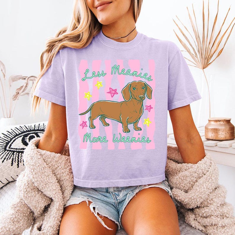 May include: A light purple short-sleeve t-shirt featuring a brown dachshund illustration in the centre. The graphic includes pink vertical stripes, colourful star accents, and the green text "Less Meanies More Weenies" printed above and below.