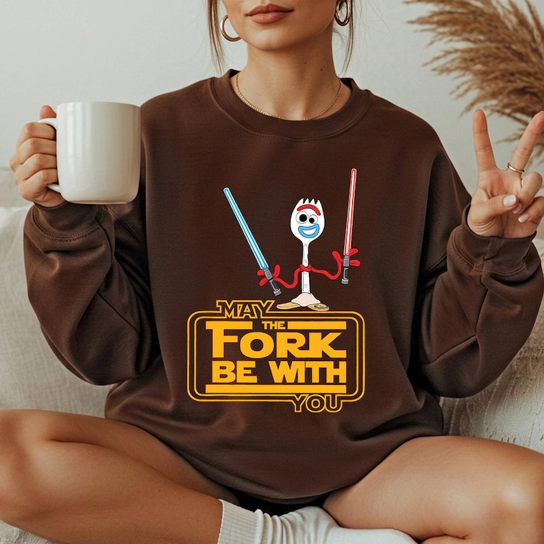 May include: A dark brown crewneck sweatshirt featuring a graphic of a white spork character holding a blue and a red lightsaber. Below the character, the yellow text reads MAY THE FORK BE WITH YOU, enclosed in a rectangular yellow outline.