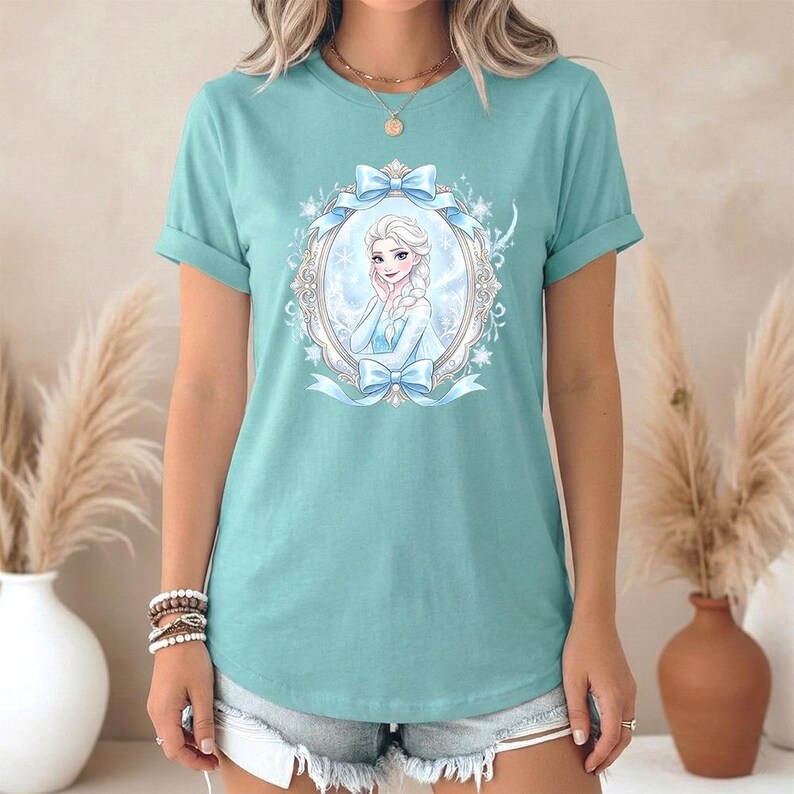 May include: A turquoise t-shirt with a framed illustration of a cartoon character with blonde braids and a blue dress. The frame is decorated with blue bows and snowflakes. The shirt has rolled-up sleeves.