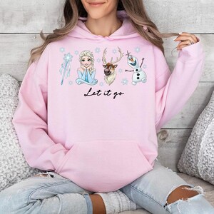 Op de afbeelding: Een lichtroze hoodie met illustraties van personages uit de film Frozen, waaronder Elsa, Olaf en Sven. De zin "Let it go" staat in zwart schrift onder de personages. De hoodie heeft een voorzak en een capuchon met trekkoord.