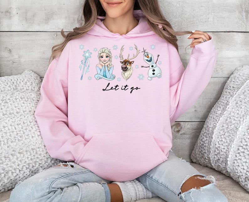 Op de afbeelding: Een lichtroze hoodie met illustraties van personages uit de film Frozen, waaronder Elsa, Olaf en Sven. De zin "Let it go" staat in zwart schrift onder de personages. De hoodie heeft een voorzak en een capuchon met trekkoord.
