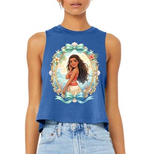 Puede incluir: Top corto azul con una ilustraci&oacute;n enmarcada de una mujer de dibujos animados con cabello largo y oscuro, vestida con un atuendo rojo y blanco, ambientada en una escena de playa tropical. El marco est&aacute; adornado con lazos, flores y conchas marinas.