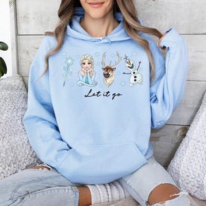 Op de afbeelding: Een lichtblauwe hoodie met illustraties van personages uit de film "Frozen", waaronder Elsa, Olaf en Sven. De zin "Let it go" is in zwart schrift geschreven. De hoodie heeft een capuchon met trekkoord en een voorzak.