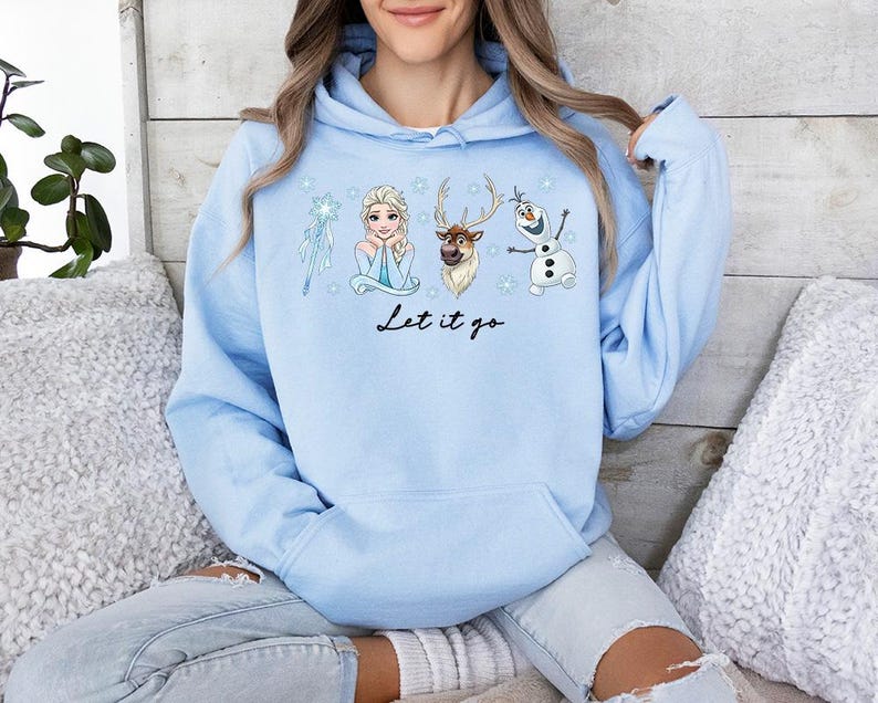 Op de afbeelding: Een lichtblauwe hoodie met illustraties van personages uit de film "Frozen", waaronder Elsa, Olaf en Sven. De zin "Let it go" is in zwart schrift geschreven. De hoodie heeft een capuchon met trekkoord en een voorzak.