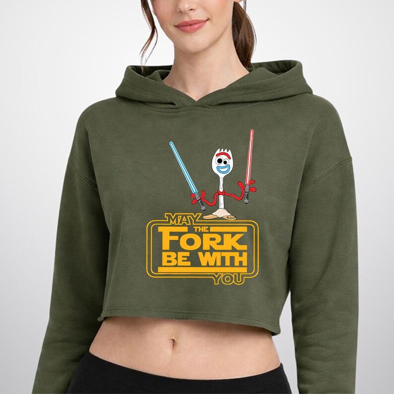 May include: An olive green cropped hoodie featuring a graphic of a white spork character with red pipe cleaner arms holding a blue and a red lightsaber. Below the character, the yellow text reads MAY THE FORK BE WITH YOU in a block style.