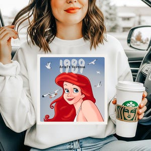 May include: White sweatshirt featuring a graphic of Ariel from The Little Mermaid, with red hair and blue eyes, and the text "1989 Ariel's Version." The design includes seagulls on a blue background. A white coffee cup with a brown sleeve is also visible.