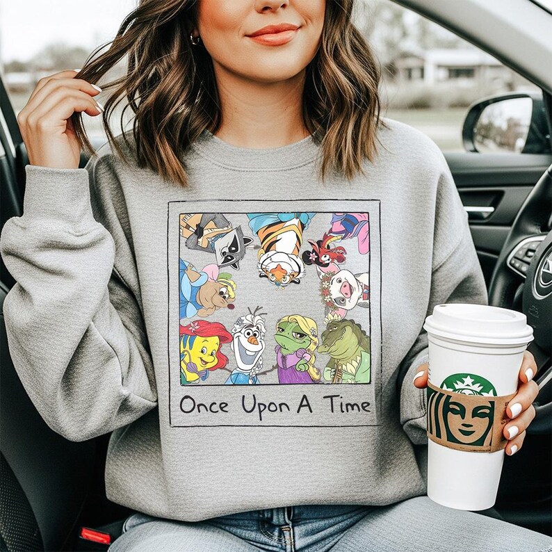 May include: A gray sweatshirt featuring a colorful graphic of cartoon characters, including a raccoon, tiger, and frog. The graphic is framed with the words "Once Upon A Time" in a rectangular box. The person is holding a white coffee cup.