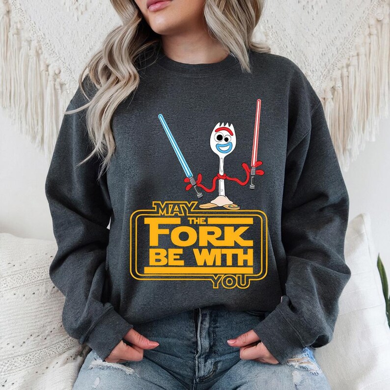 May include: A dark grey crewneck sweatshirt featuring a graphic of the character Forky holding a blue and a red lightsaber. Below the character, the yellow text reads: MAY THE FORK BE WITH YOU, styled in a classic science fiction movie font.