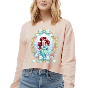 Puede incluir: Una sudadera corta de color rosa claro con una ilustraci&oacute;n enmarcada de una sirena con cabello rojo y un vestido turquesa. El marco es dorado con lazos turquesa en la parte superior e inferior. La sirena est&aacute; rodeada de conchas marinas y plantas acu&aacute;ticas.