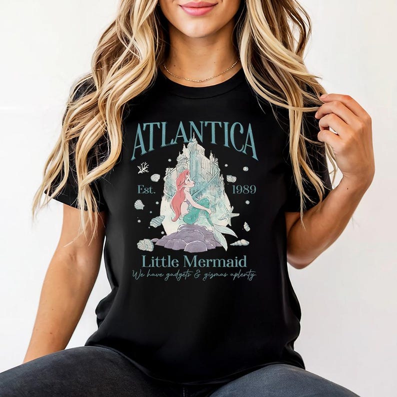 Puede incluir: Camiseta negra con un gr&aacute;fico turquesa. El dise&ntilde;o incluye la palabra "Atlantica", una ilustraci&oacute;n de una sirena y un castillo, y el texto "Est. 1989" y "Little Mermaid". La camiseta tambi&eacute;n tiene el texto "We have gadgets & gizmos aplenty."