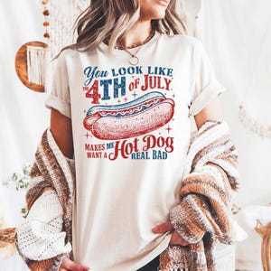 Peut inclure: T-shirt crème avec un motif graphique de hot-dog et du texte. Le texte dit "You look like the 4th of July makes me want a hot dog real bad" en rouge et bleu. Le motif du hot-dog est rouge et blanc.