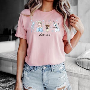 May include: A light pink t-shirt featuring illustrations of characters from the movie Frozen, including Elsa, Olaf, and Sven, with the text "Let it go" in a script font. The shirt is made of a soft material.