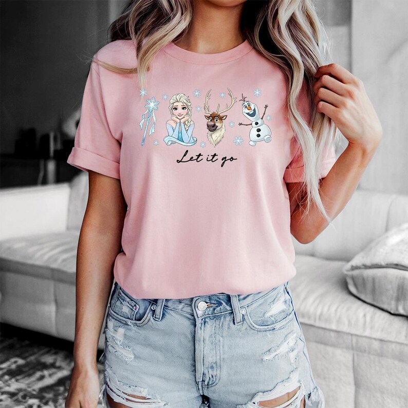 May include: A light pink t-shirt featuring illustrations of characters from the movie Frozen, including Elsa, Olaf, and Sven, with the text "Let it go" in a script font. The shirt is made of a soft material.