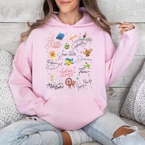 May include: A light pink hooded sweatshirt featuring a central graphic print of various character names in cursive script, including Belle, Cinderella, Snow White, Jasmine, Rapunzel, Ariel, Fiona, and Sleeping Beauty, alongside small colorful icons.