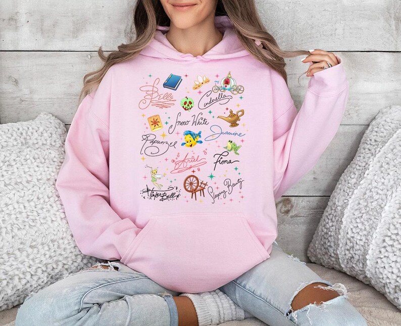 May include: A light pink hooded sweatshirt featuring a central graphic print of various character names in cursive script, including Belle, Cinderella, Snow White, Jasmine, Rapunzel, Ariel, Fiona, and Sleeping Beauty, alongside small colorful icons.
