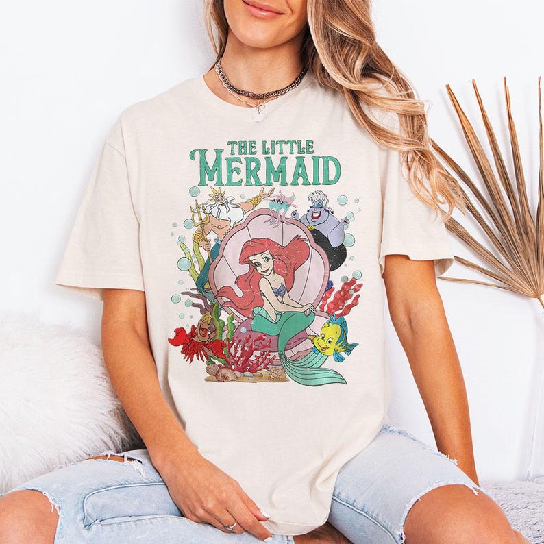 May include: A cream-coloured short-sleeve t-shirt featuring a graphic print of The Little Mermaid characters including Ariel, King Triton, Ursula, Sebastian, and Flounder. The design includes the text THE LITTLE MERMAID in green lettering.