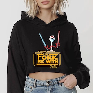 May include: A black cropped hoodie featuring a graphic of a white plastic fork character with red pipe cleaner arms holding a blue and a red lightsaber. Below the character, the yellow text reads: MAY THE FORK BE WITH YOU.
