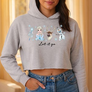 May include: A light grey cropped hoodie with a graphic of Elsa, Olaf, Sven, and a snowflake wand. The text "Let it go" is printed below the characters. The hoodie has a drawstring hood and long sleeves.