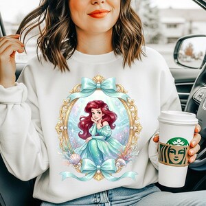 May include: White sweatshirt featuring a cartoon illustration of a mermaid in a gold frame with blue bows. The mermaid has red hair and is wearing a teal dress. The design includes seashells and aquatic elements.