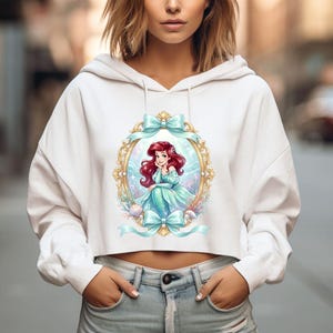 Op de afbeelding: Witte cropped hoodie met een kleurrijke illustratie van een zeemeermin in een gouden lijst met blauwe strikken. De zeemeermin heeft rood haar en draagt een turquoise jurk. Het ontwerp bevat schelpen en waterplanten.