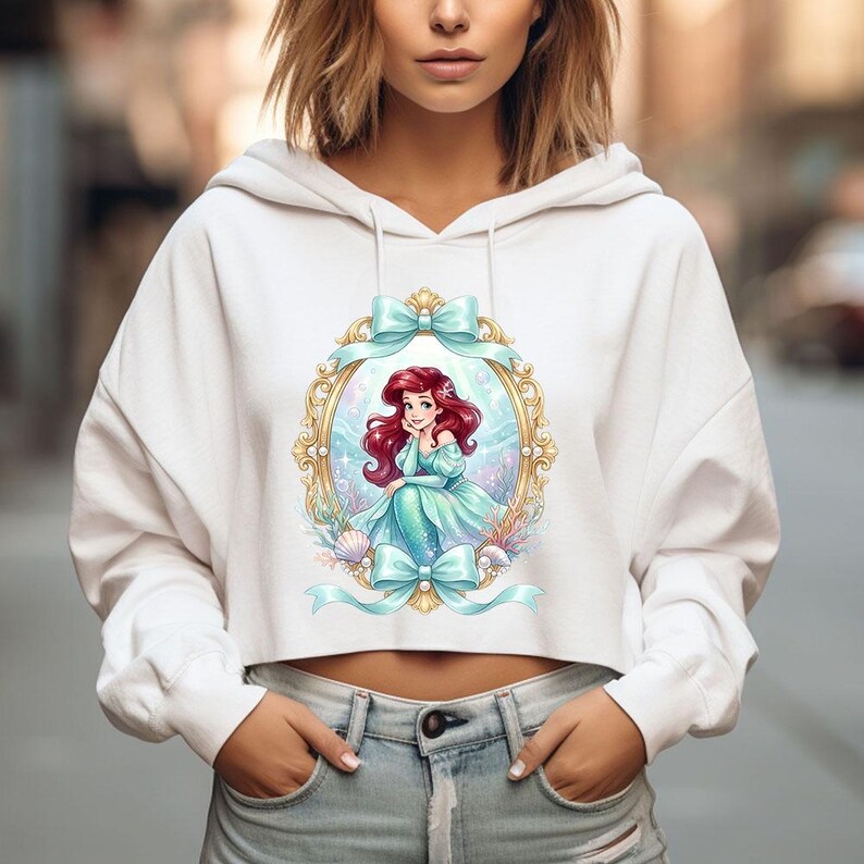 Op de afbeelding: Witte cropped hoodie met een kleurrijke illustratie van een zeemeermin in een gouden lijst met blauwe strikken. De zeemeermin heeft rood haar en draagt een turquoise jurk. Het ontwerp bevat schelpen en waterplanten.