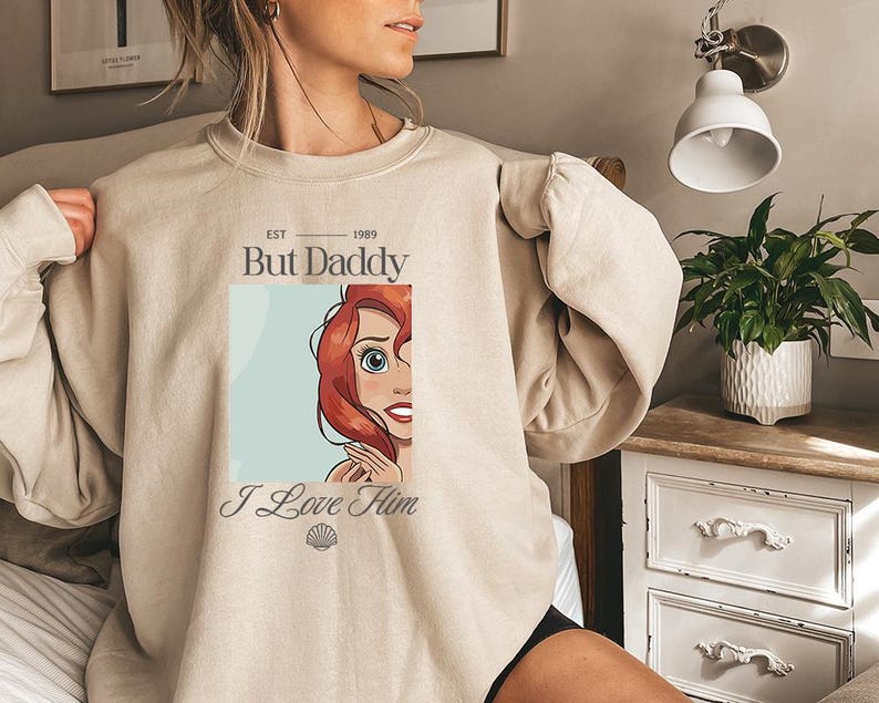 May include: Beige sweatshirt featuring a graphic of a cartoon character with red hair and blue eyes. The text "But Daddy" and "I Love Him" are printed on the front. The sweatshirt has long sleeves and a crew neck.