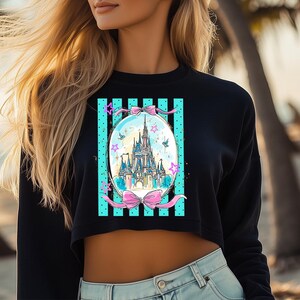 May include: Black cropped sweatshirt featuring a colorful illustration of a fairytale castle inside an oval frame. The frame has a teal background with vertical black stripes and pink bows. The design includes stars and birds.