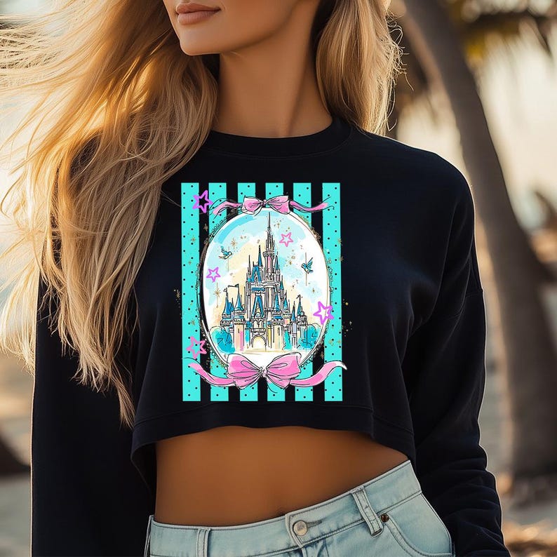 May include: Black cropped sweatshirt featuring a colorful illustration of a fairytale castle inside an oval frame. The frame has a teal background with vertical black stripes and pink bows. The design includes stars and birds.