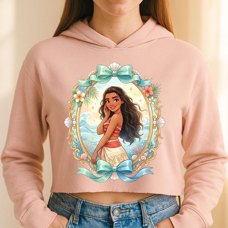 K&ouml;nnte beinhalten: Ein hellrosa Crop-Hoodie mit einer farbenfrohen Illustration einer Frau in einem ovalen Rahmen. Der Rahmen enth&auml;lt ein blaues Band, Blumen und Muscheln. Die Frau hat lange dunkle Haare und tr&auml;gt ein rot-wei&szlig;es Outfit.