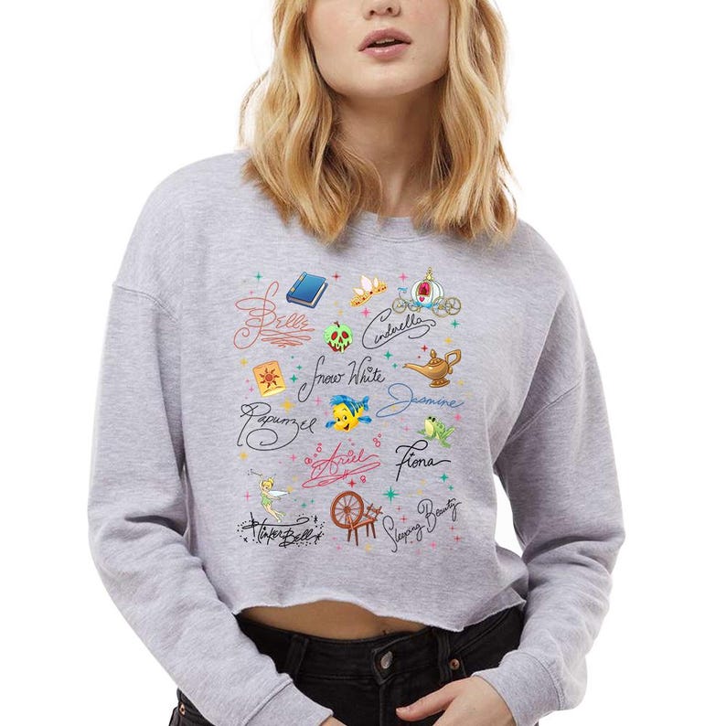May include: A light grey cropped sweatshirt featuring a graphic print of various character names in cursive script, including Belle, Cinderella, Snow White, Jasmine, Rapunzel, Ariel, Fiona, Tinker Bell, and Sleeping Beauty, alongside small colourful icons.