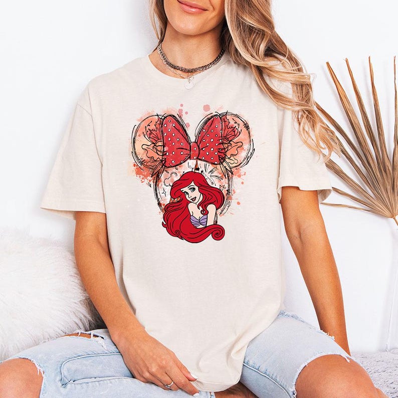 Puede incluir: Camiseta color crema con un gr&aacute;fico de Ariel de La Sirenita. El dise&ntilde;o incluye un lazo rojo con lunares, orejas de Mickey Mouse y detalles de acuarela. La camiseta es de corte holgado.