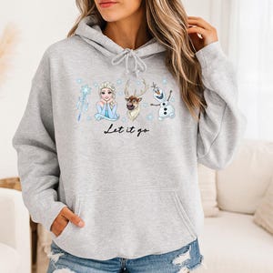 Op de afbeelding: Lichtgrijze hoodie met een afbeelding van Elsa, een rendier en Olaf uit Frozen, met de tekst "Let it go". Het ontwerp bevat sneeuwvlokken en een toverstaf. De hoodie heeft een capuchon met trekkoord en een voorzak.