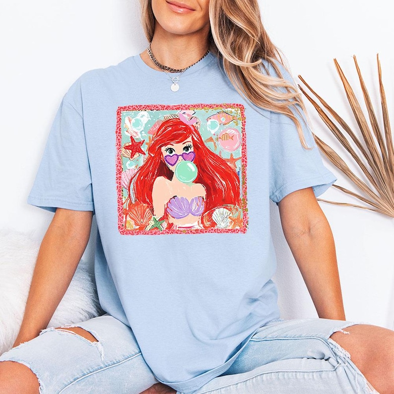 May include: Light blue t-shirt featuring a colorful graphic of a cartoon mermaid with red hair, heart-shaped sunglasses, and a bubble. The graphic is surrounded by sea-themed elements like starfish and seashells.
