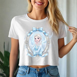 Pode incluir: Top curto branco com uma ilustra&ccedil;&atilde;o emoldurada de uma personagem de desenho animado com cabelo loiro e um vestido azul. A moldura &eacute; decorada com la&ccedil;os azuis e flocos de neve. A camisa &eacute; feita de um material macio e confort&aacute;vel.