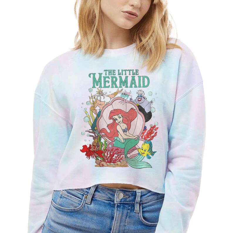 May include: A long-sleeve crop sweatshirt in a pastel tie-dye pattern featuring a graphic print of The Little Mermaid characters including Ariel, King Triton, Ursula, Sebastian, and Flounder with the text THE LITTLE MERMAID printed above them.