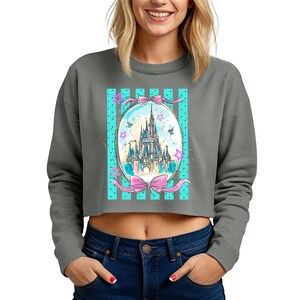 May include: Gray cropped sweatshirt featuring a colorful illustration of a fairytale castle within an oval frame. The frame is decorated with pink bows, stars, and teal stripes. The sweatshirt has long sleeves and a crew neck.