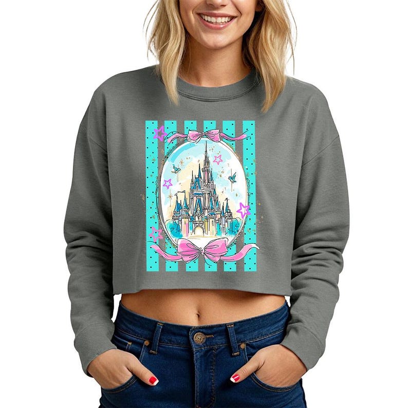 May include: Gray cropped sweatshirt featuring a colorful illustration of a fairytale castle within an oval frame. The frame is decorated with pink bows, stars, and teal stripes. The sweatshirt has long sleeves and a crew neck.
