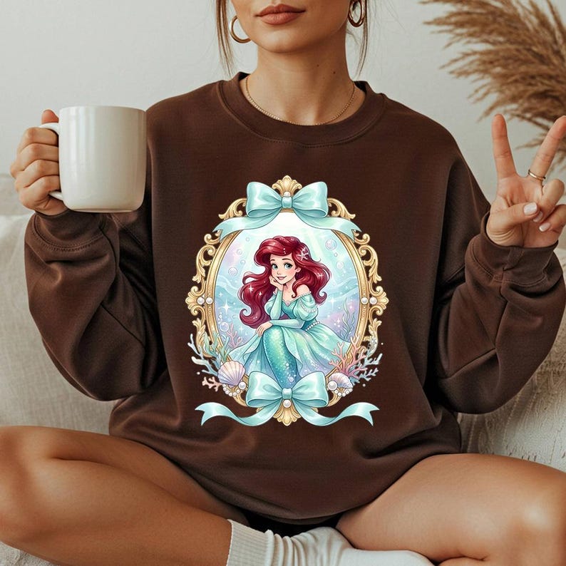 May include: Brown sweatshirt featuring a framed illustration of a mermaid with red hair and a teal dress. The frame is gold with teal bows at the top and bottom. The mermaid is surrounded by seashells and aquatic plants.