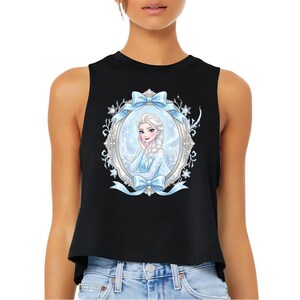 Pu&ograve; includere: Top corto senza maniche nero con un'illustrazione incorniciata di un personaggio dei cartoni animati con i capelli biondi e un vestito blu. L'illustrazione &egrave; circondata da un fiocco azzurro e un motivo floreale.