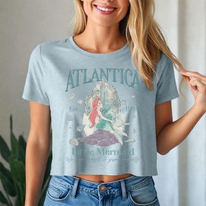 May include: Light blue crop top featuring a graphic of the Little Mermaid. The design includes the word "Atlantica" above a castle illustration, the mermaid, and the text "Est. 1989". The shirt is made of a soft material.