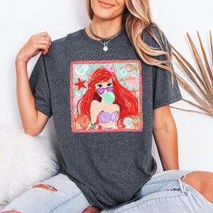 May include: Dark gray t-shirt featuring a colorful graphic of a mermaid with red hair, heart-shaped sunglasses, and a bubble. The graphic is surrounded by seashells, starfish, and fish. The mermaid is wearing a purple seashell bra.