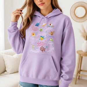 May include: A light purple hooded sweatshirt featuring a front graphic print of various animated character icons and names in cursive script, including a book, a crown, a carriage, a lamp, a fish, a spinning wheel, and a fairy, with a front pouch pocket.