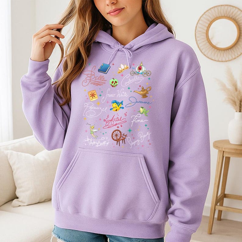 May include: A light purple hooded sweatshirt featuring a front graphic print of various animated character icons and names in cursive script, including a book, a crown, a carriage, a lamp, a fish, a spinning wheel, and a fairy, with a front pouch pocket.