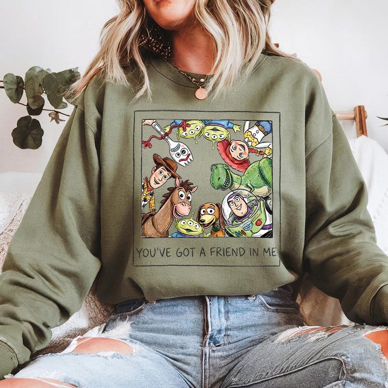 May include: An olive green sweatshirt with a framed graphic of Toy Story characters, including Woody and Buzz Lightyear. The graphic is surrounded by a black border. The text "YOU'VE GOT A FRIEND IN ME" is printed below the graphic.